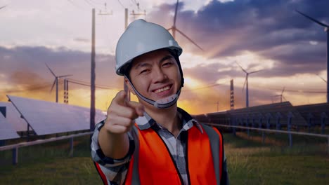 close up of asian male engineer with safety helmet smiling and touching his chest then pointing at you while standing with solar panel and wind turbines