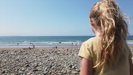 Niña-Rubia-De-Siete-Años-Con-Camiseta-Amarilla-Subiendo-Un-Banco-De-Guijarros-Para-Revelar-Una-Concurrida-Playa,-Mar---Horizonte-En-Un-Claro-Día-De-Verano