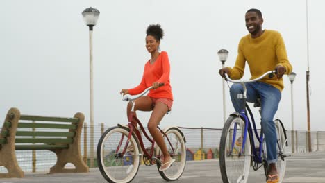 Front-view-of-black-young-couple-riding-bicycle-on-promenade-at-beach-on-a-sunny-day-4k