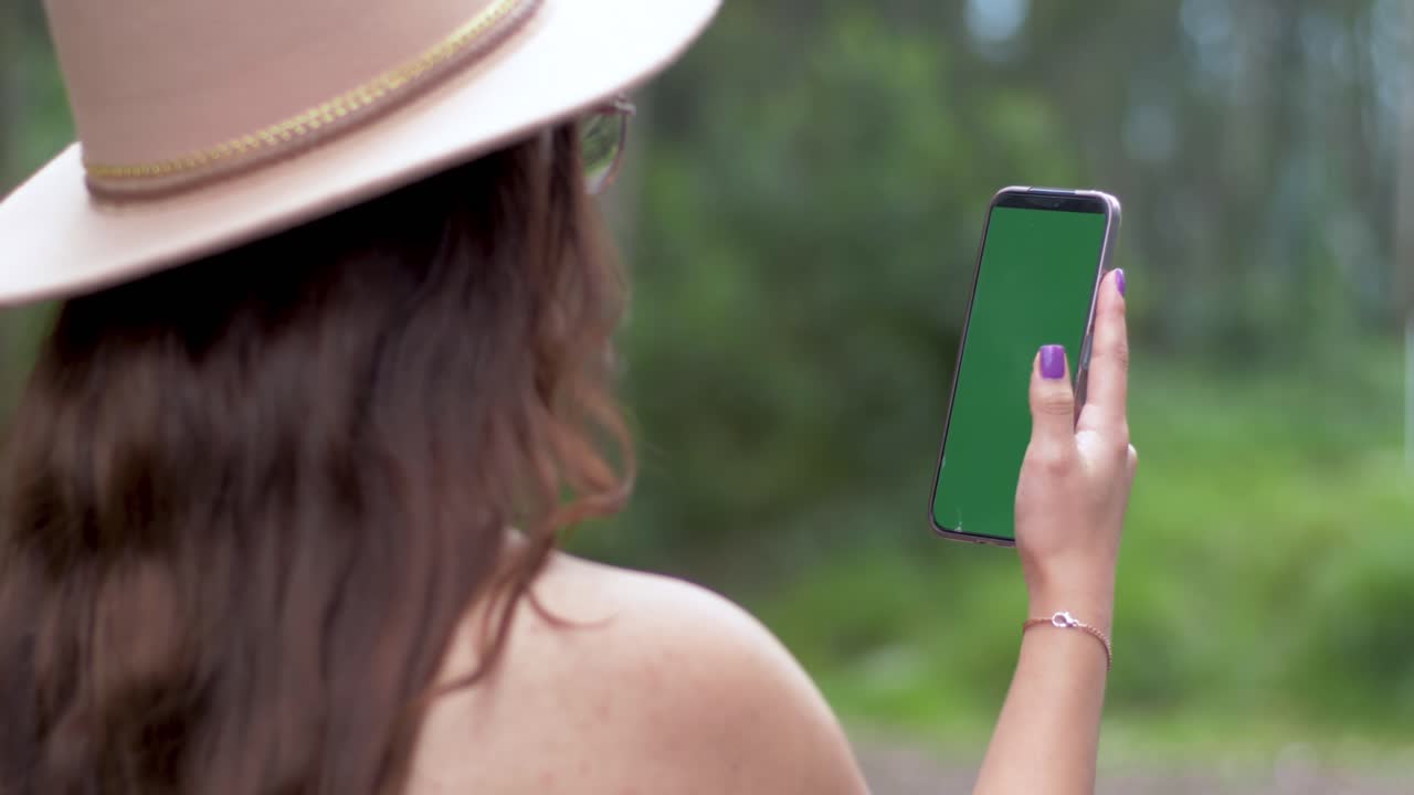 una chica con el cabello abierto, con un sombrero y gafas, se involucra con su teléfono inteligente contra un telón de fondo de árboles verdes y hierba