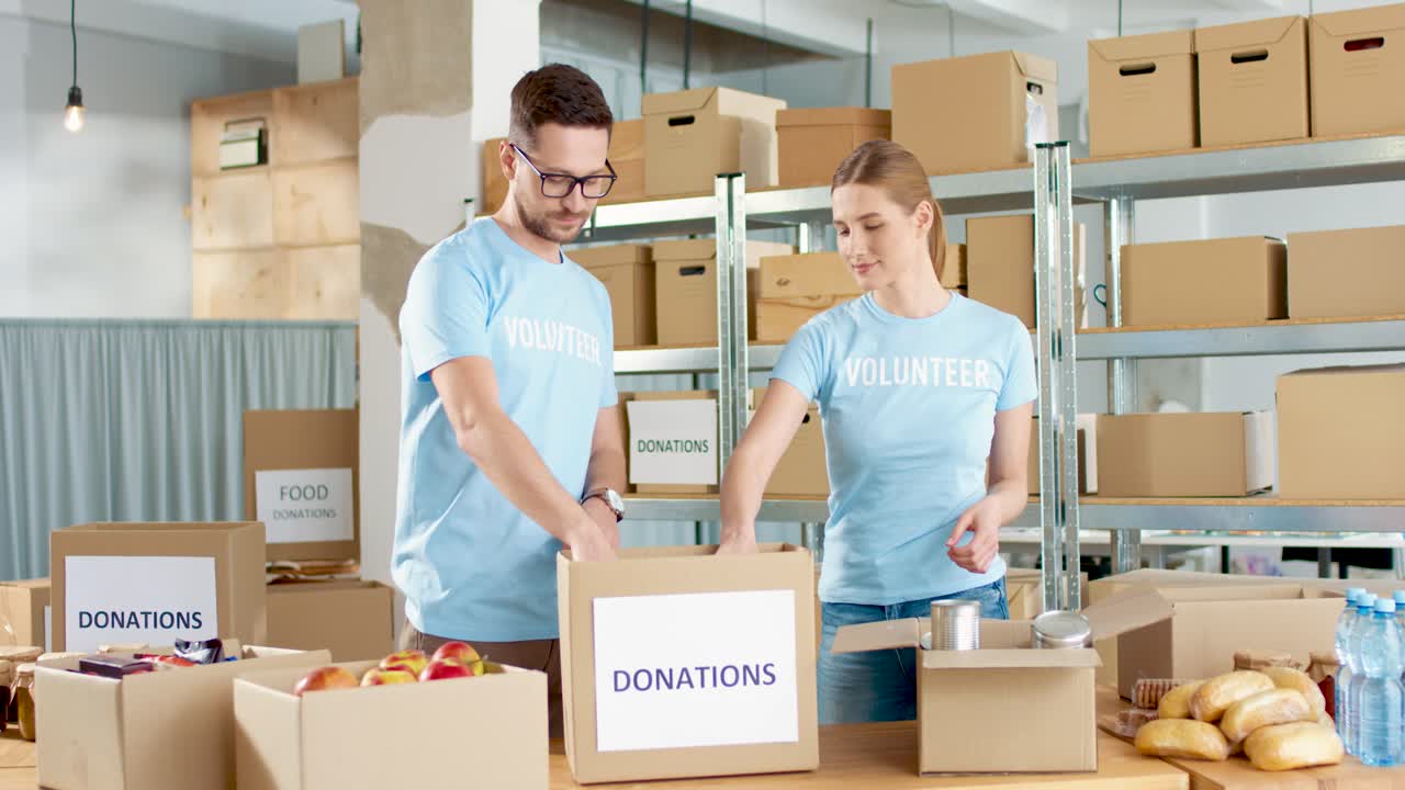 jóvenes voluntarios caucásicos, hombres y mujeres, empacando una caja con comida y luego sonriendo a la cámara en un almacén de caridad