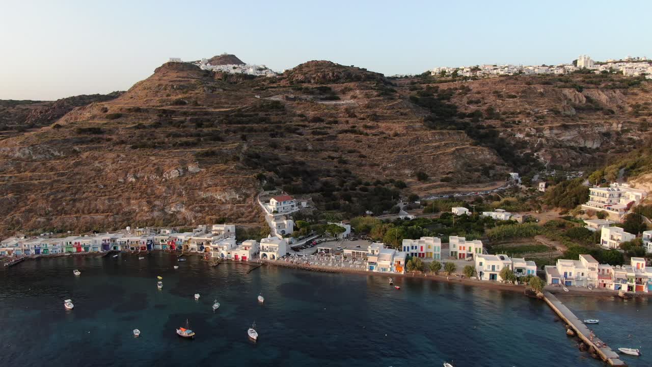 drone vista en grecia vertical ascender sobre una pequeña ciudad con casas blancas y puertas de colores junto al mar en frente de una montaña en milos al atardecer