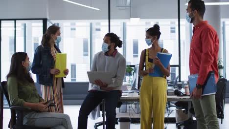 Diverse-group-of-work-colleagues-wearing-face-mask-holding-laptop,-tablet-and-documents-talking