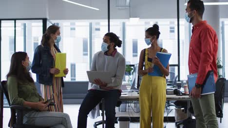 grupo diverso de colegas de trabajo con máscara facial sosteniendo una computadora portátil, tableta y documentos hablando