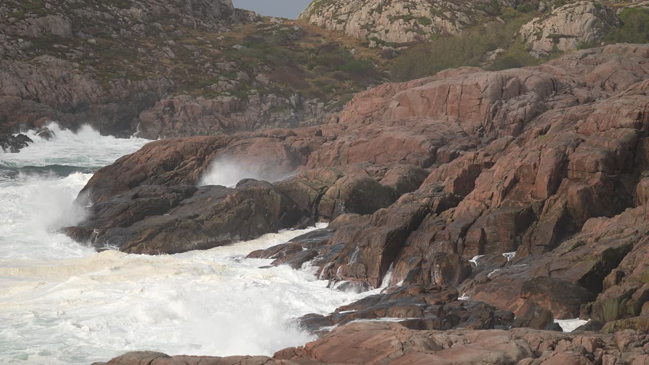 Turbulent waves collide with rocky cliffs at a coastal location. The scene captures the natural beauty and power of the ocean under a cloudy sky with rolling hills in the background