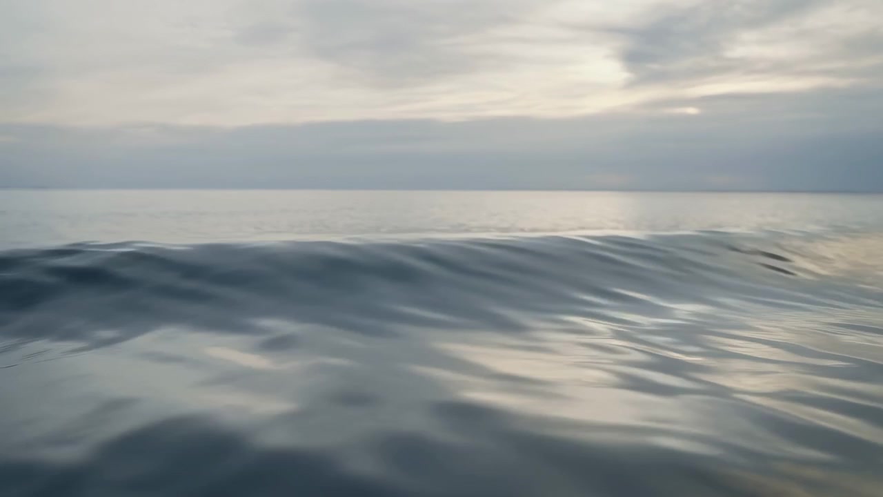 Low-angle seascape showing gentle swell and fine ripple movement across smooth ocean water, with silvery reflections, muted gray tones, layered clouds, and quiet distant horizon, with ocean, sea