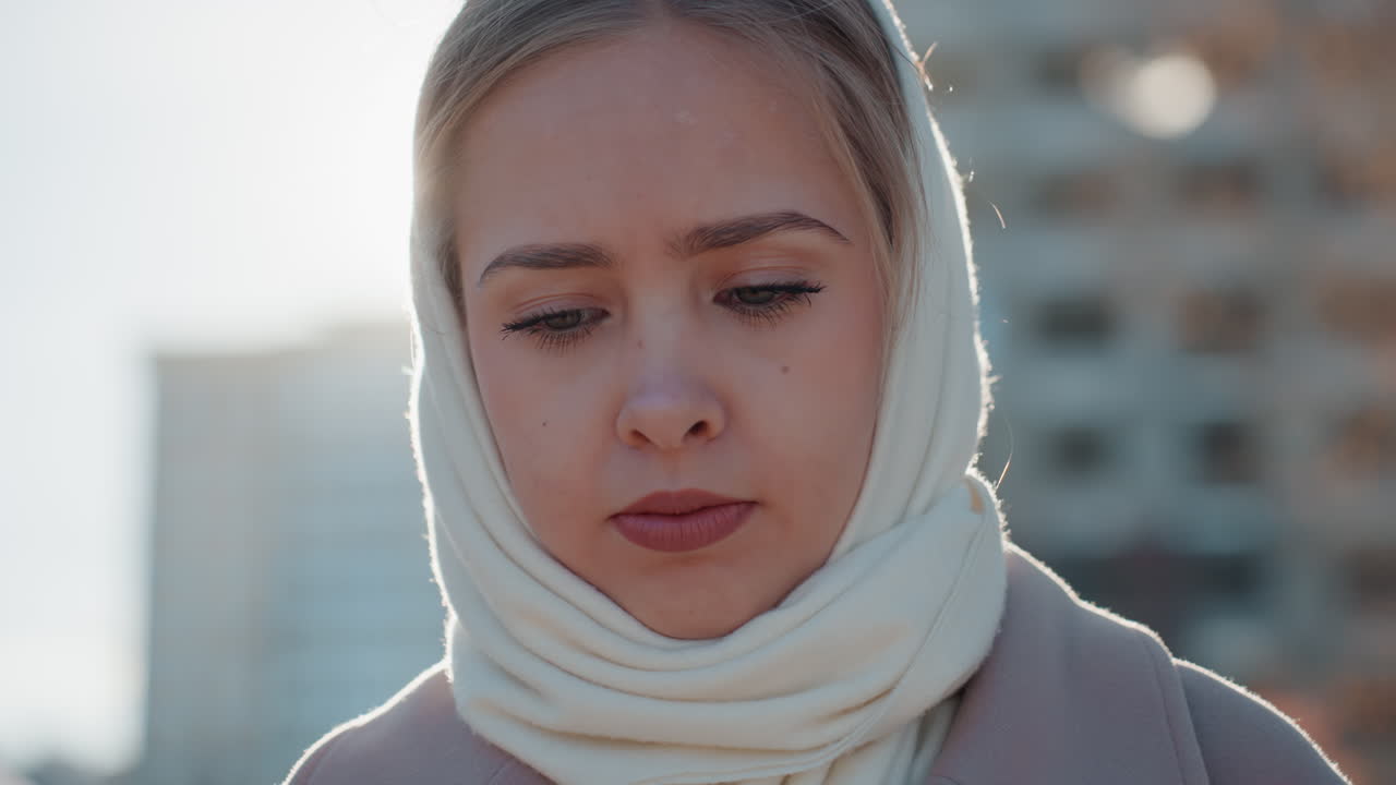 Yansıtıcı yakın çekimde hüzünlü bakışlı beyaz tenli bir kadın; arkadan aydınlatmalı şehir arka planı ve yumuşak bokeh efektiyle. Yüzünü çevreleyen krem rengi eşarp, soluk sonbahar tonları ve sinematik bir havayla sessiz, düşünceli bir ruh hali yansıtıyor.