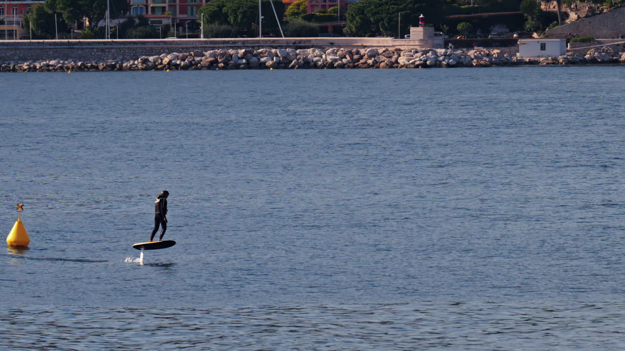 Villefranche sur Mer, France - March 24, 2025: Man surf foiling alone on the sea with the buildings of the city on the background