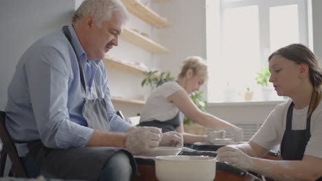 Medium-shot-of-middle-aged-ceramic-artist-teaching-group-elderly-Caucasian-woman-and-senior-man-how-to-wedge-clay-sitting-at-desk-in-art-studio.-People-enjoying-talking-at-work