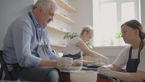 disparo medio de un artista de cerámica de mediana edad que enseña a un grupo de ancianos, una mujer caucásica y un hombre mayor cómo hacer arcilla sentados en un escritorio en un estudio de arte. personas que disfrutan hablando en el trabajo