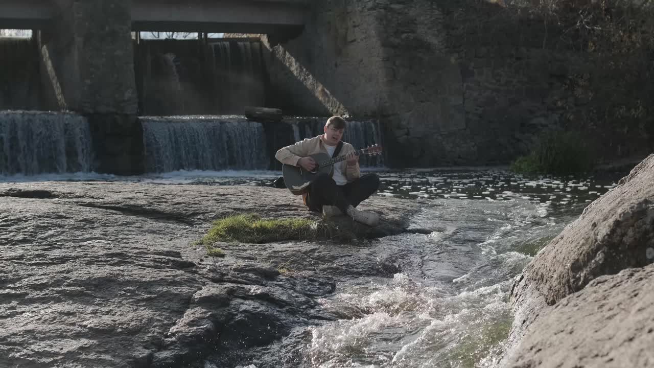 joven tocando la guitarra sentado en la orilla de un río de montaña sobre un fondo de rocas. concepto de libertad lugar de relajación.