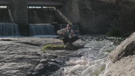 young man playing guitar sitting on the bank of a mountain river on a background of rocks. concept of freedom relaxation. place