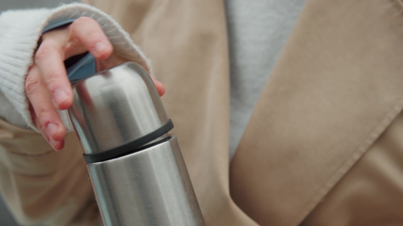 Primo piano di una donna che apre e chiude il coperchio di un thermos in acciaio inossidabile mentre indossa un cappotto beige. L'azione è catturata in dettaglio mostrando l'interazione con oggetti metallici in un contesto urbano esterno.