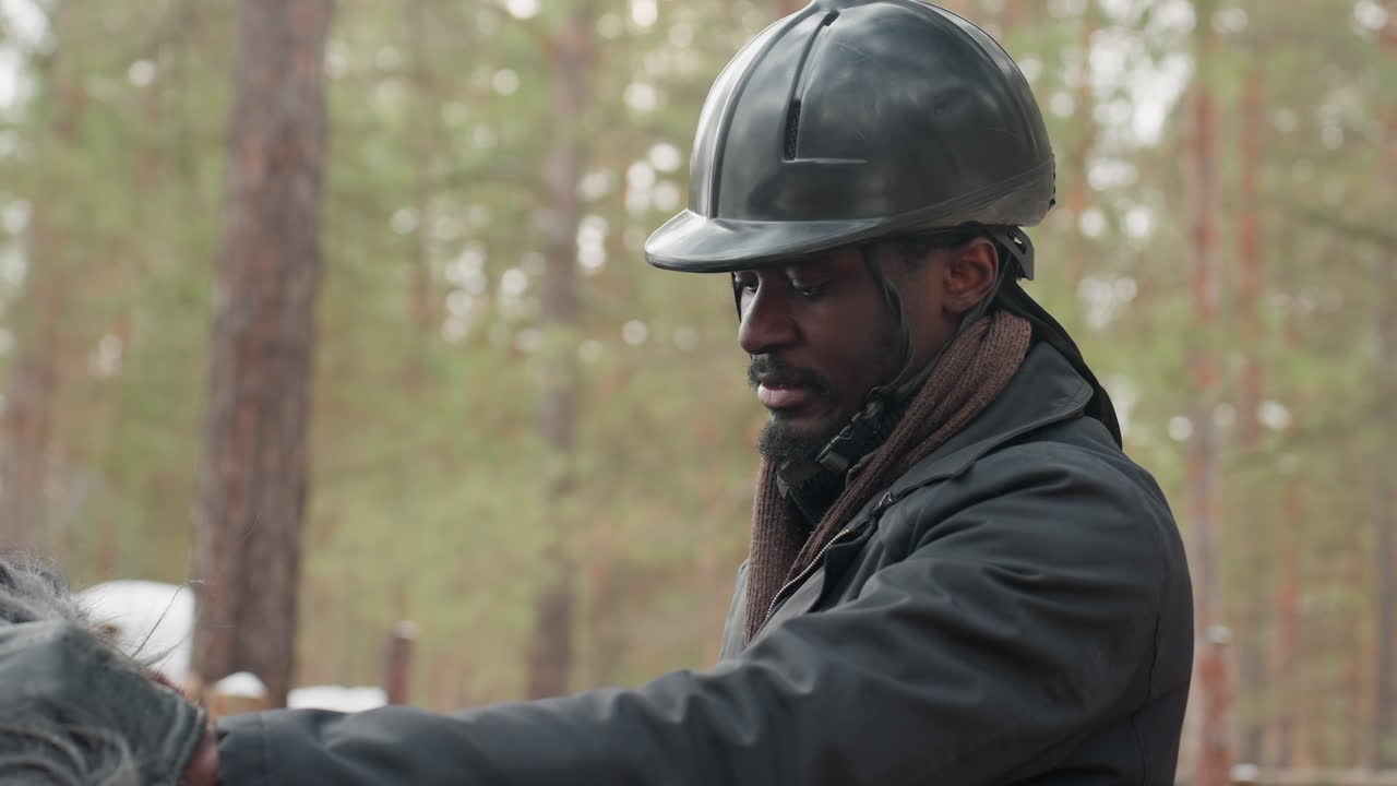 Фотография темнокожего инструктора в профиль, едущего верхом по лесу, сохраняющего устойчивую позу и внимательное выражение лица, в шлеме и плотной куртке на фоне приглушенного лесного пейзажа, руки на поводьях уверенно направляют лошадь