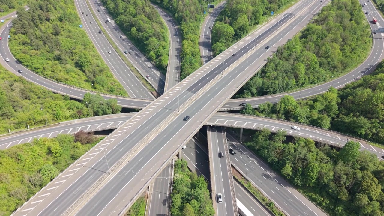 Aerial perspective of a complex highway interchange. Cars and trucks drive on multiple roads and bridges surrounded by green trees. Daytime traffic moves smoothly through the multilevel network.