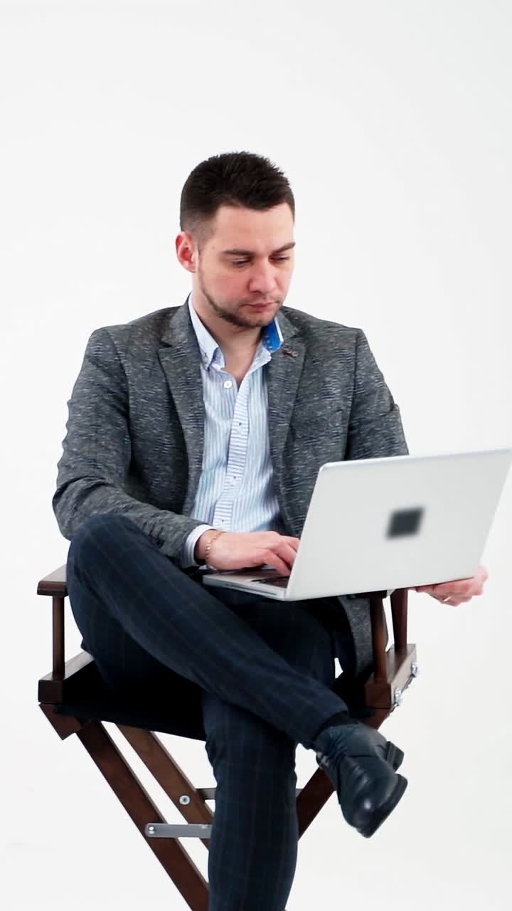 Young man sitting in a chair with laptop. Handsome businessman in stylish costume working on a modern computer, isolated in studio. Freelance work. Vertical video