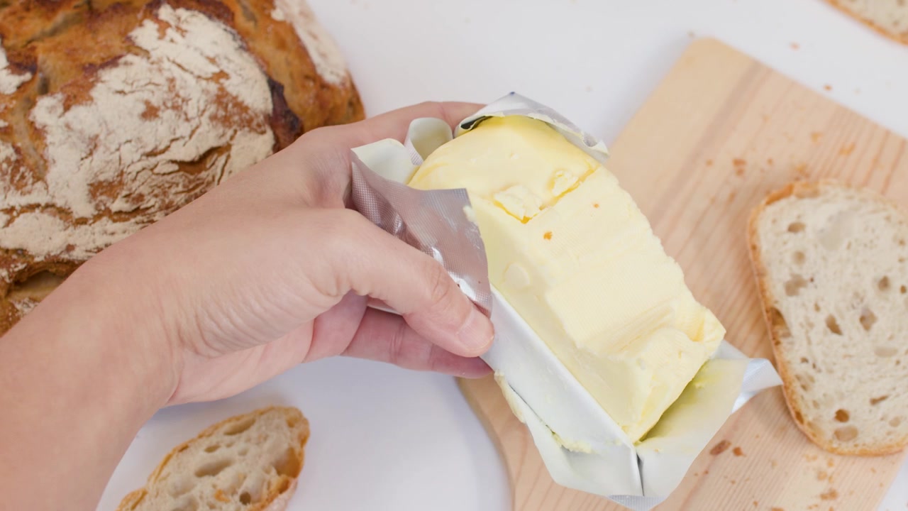 Top view of hands arranging sourdough bread and butter on a wooden board against white.