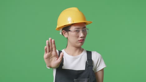 close up of asian woman worker wearing goggles and safety helmet looking at camera and disapproving with no hand sign while standing in the green screen background studio