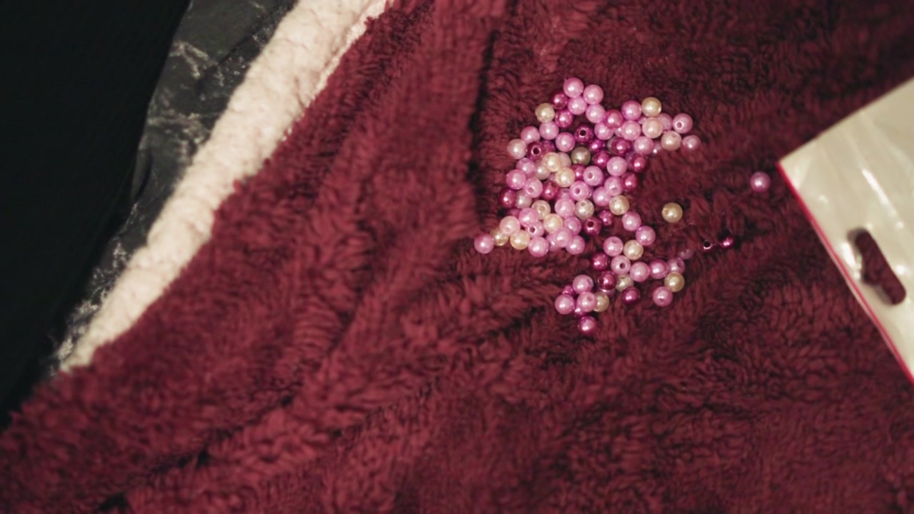 Perline rosa su asciugamano bordeaux sparse come gettoni oracolari, superficie morbida e testurizzata e ombre delicate, primo piano con atmosfera mistica che suggerisce divinazione, fascino talismanico e preveggenza profetica.