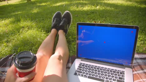 closeup portrait of young pretty caucasian female legs girl sitting on rug holding coffee using laptop with blue chroma screen in park outdoors