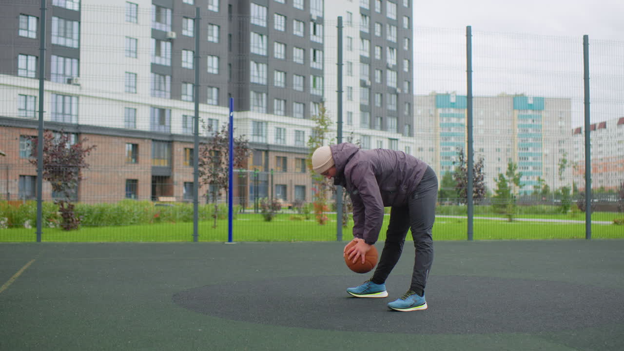 Elderly white man picking up basketball on city playground, adjusting grip and bending with careful posture, sneakers on asphalt near apartment skyline, solo training session with deliberate technique