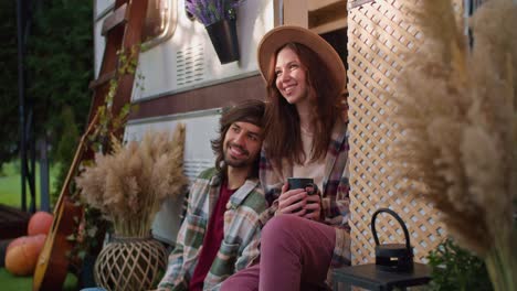 Happy-brunette-girl-in-a-light-brown-black-mug-with-a-hot-drink-in-her-hands-and-communicates-with-her-brunette-boyfriend-in-a-green-checkered-shirt-sitting-near-a-decorated-trailer-in-a-camp-during-a-picnic-outside-the-city-in-the-summer