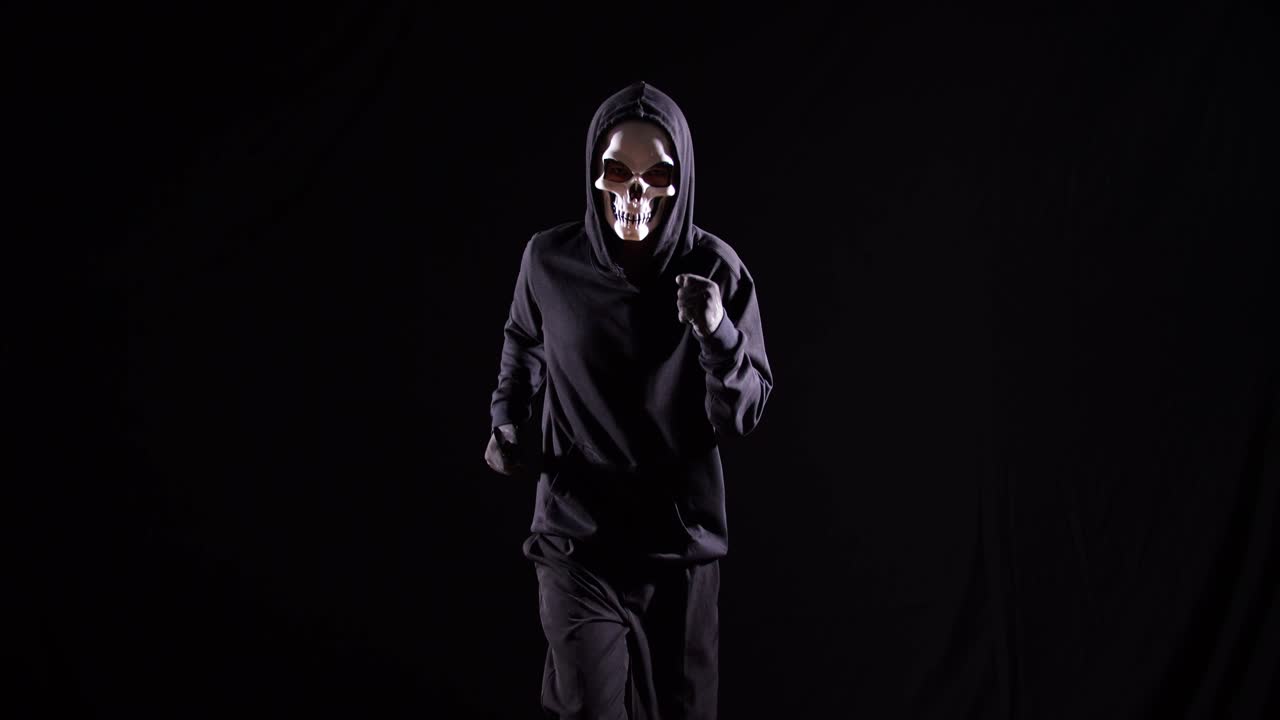 hombre aterrador en la sudadera con capucha usando una máscara de halloween sosteniendo un cuchillo y corriendo sobre el fondo negro
