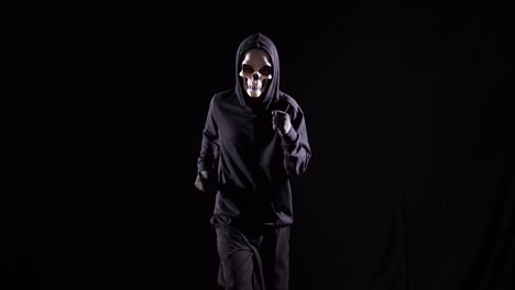 hombre aterrador en la sudadera con capucha usando una máscara de halloween sosteniendo un cuchillo y corriendo sobre el fondo negro
