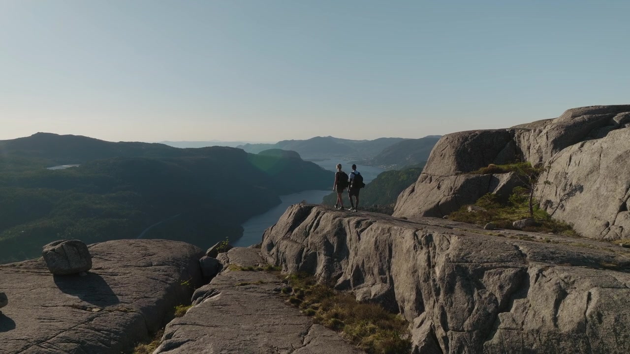 Una coppia di escursionisti cammina su una lastra di granito verso un punto panoramico spettacolare con vista sul Lysefjord durante una soleggiata giornata estiva nordica sull'escursione Hatten in Norvegia.