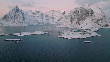 Vista-Aérea-Descendente-De-Las-Nevadas-Montañas-Moskenes-Hasta-El-Pintoresco-Pueblo-Pesquero-De-Reine-Al-Amanecer