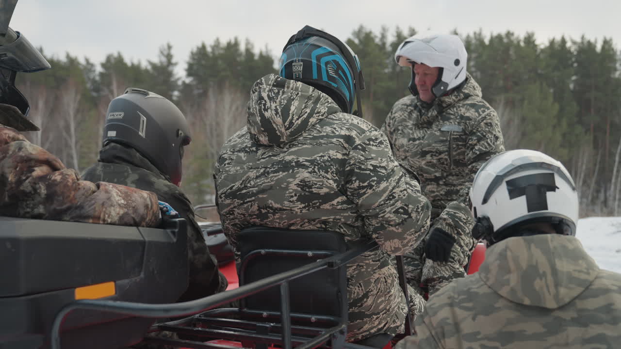 Group prepping snowmobile in snowy forest with helmeted riders checking gear, loading sled and coordinating route before offroad ride camo jackets, throttle check, friendly briefing and mechanical