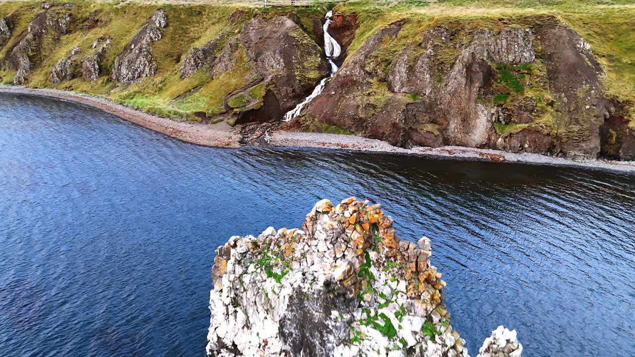 Little waterfall running among the rocks. Drone rising over the mountains covered with moss. A piece of rocks sticks out of the water.