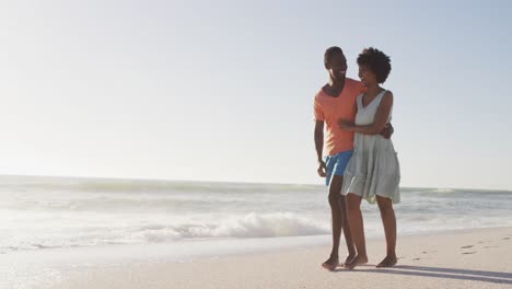 Smiling-african-american-couple-embracing-and-walking-on-sunny-beach