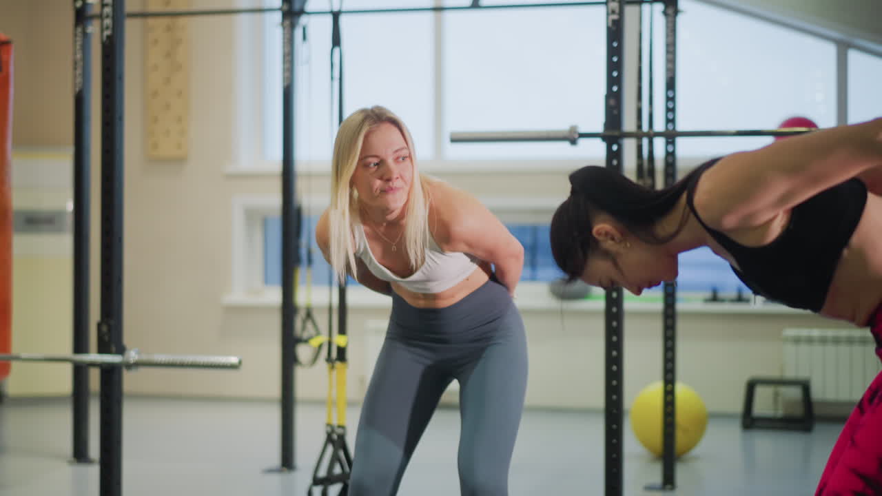 two women in gym stretching, trainer guiding client through triceps extension and posture correction, bright studio with racks and medicine balls, focused warmup, casual athletic attire, supportive