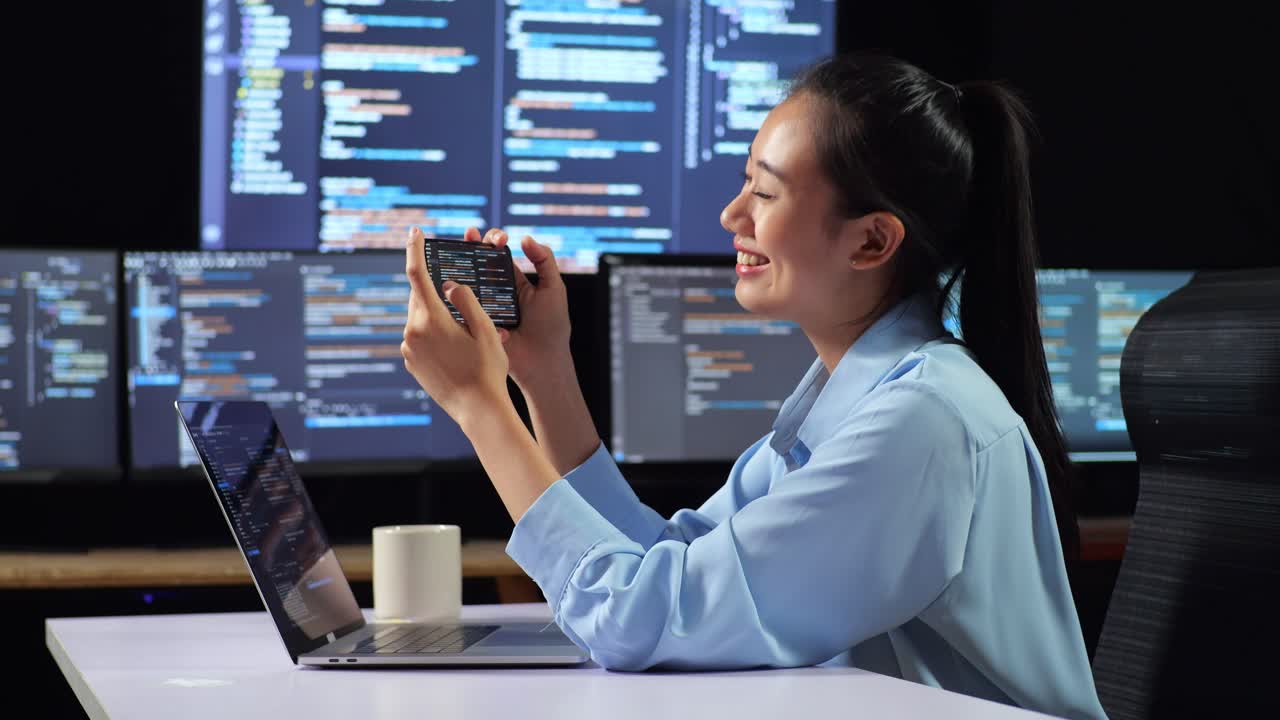 아시아 여성 프로그래머가 스마트 폰을 보고 노트북으로 코드를 작성하는 동안 성공을 축하하는 측면 뷰 여러 모니터를 사용하여 사무실의 데스크에서 데이터베이스를 보여줍니다.