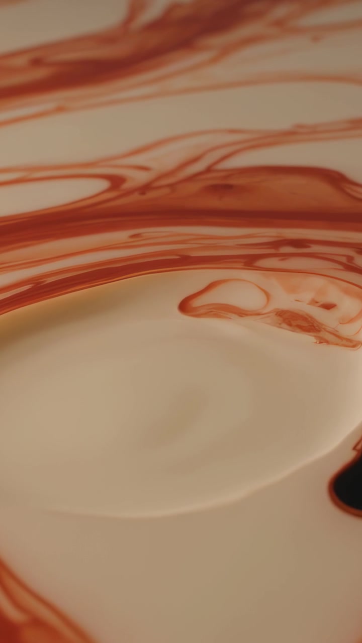 Video vertical: Gota marrón cayendo y extendiéndose sobre leche en un plato blanco, formando un charco a la derecha.