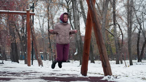 dama vestida cálidamente con chaqueta de invierno y pantalones de color marrón disfrutando de un columpio en el patio de recreo en un día de invierno nevado, sosteniendo una cuerda de capucha con árboles desnudos y suelo cubierto de nieve en el fondo