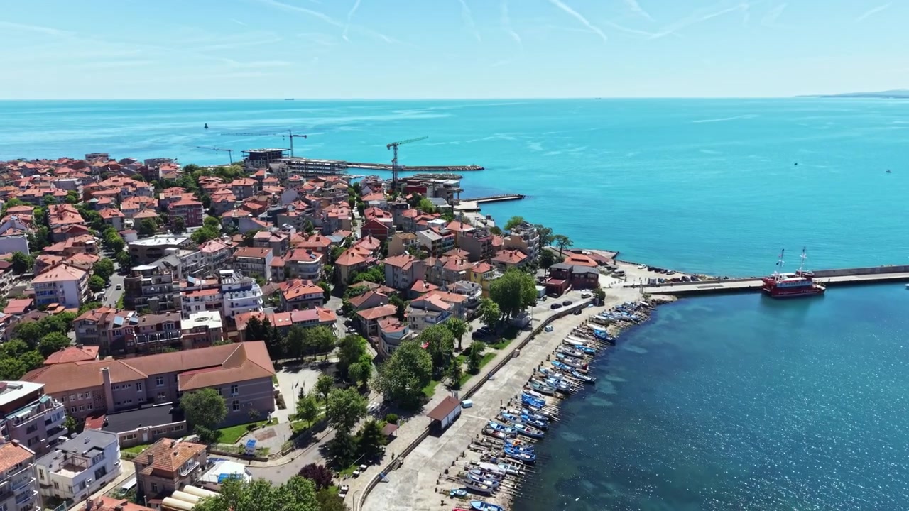 Güneşli bir günde, liman boyunca demirlemiş tekneler ve mavi denizin yakınındaki yamaçta evler bulunan bir sahil kasabası manzarası.
