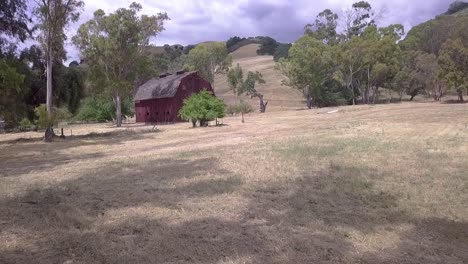 Beautiful,-rustic-red-barn-in-the-countryside,-drone-shot-moving-towards-then-over-barn-to-reveal-green-tress-and-golden-hills-of-California,-and-blue-sky-with-clouds