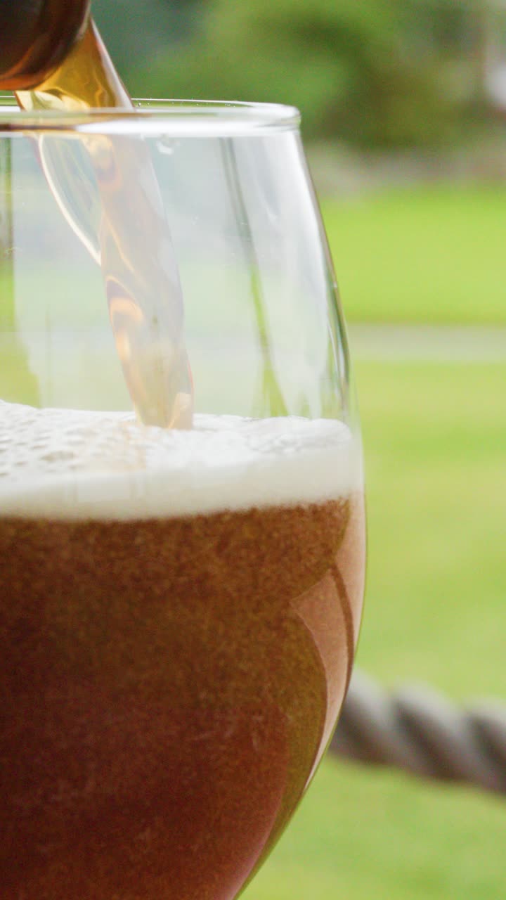Birra ambrata viene versata da una bottiglia in un bicchiere trasparente, formando una schiuma densa, con uno sfondo di giardino verde e luce naturale del giorno