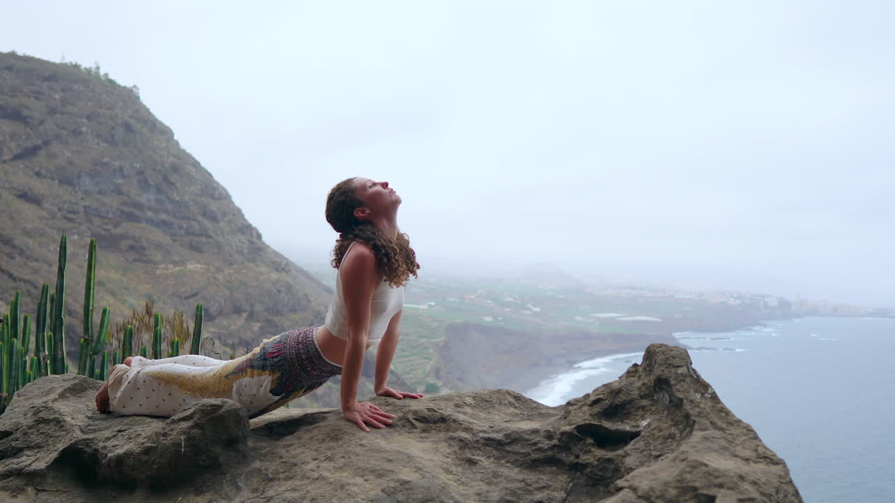 The woman sits on a cliff, in a dog pose, gazing at the ocean, taking in the sea air as part of her yoga journey through the islands