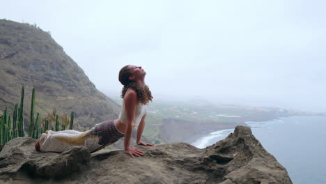 The-woman-sits-on-a-cliff,-in-a-dog-pose,-gazing-at-the-ocean,-taking-in-the-sea-air-as-part-of-her-yoga-journey-through-the-islands