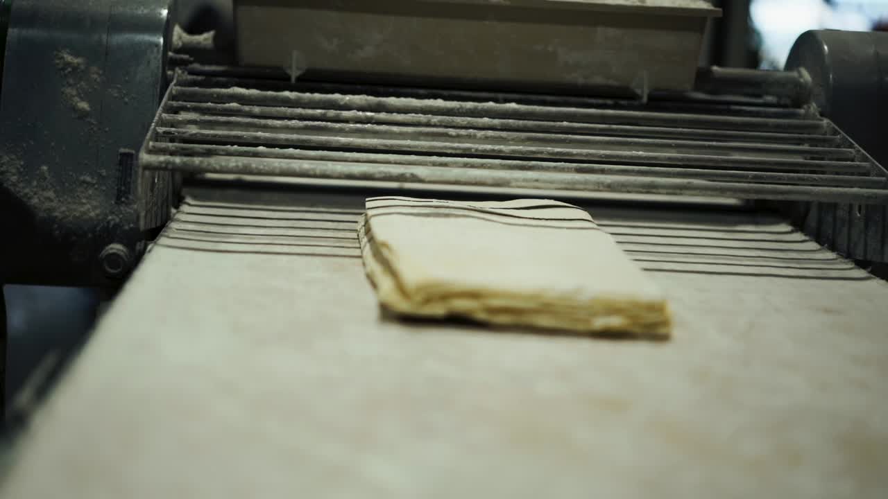 Medium shot of slab roller croissant dough machine rolling sheets of laminated croissant dough, focus pulling from machine to pastry