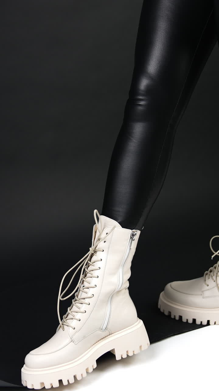 Woman in black leather leggings wears white middle-length boots with tractor soles. Model posing in studio. Vertical video