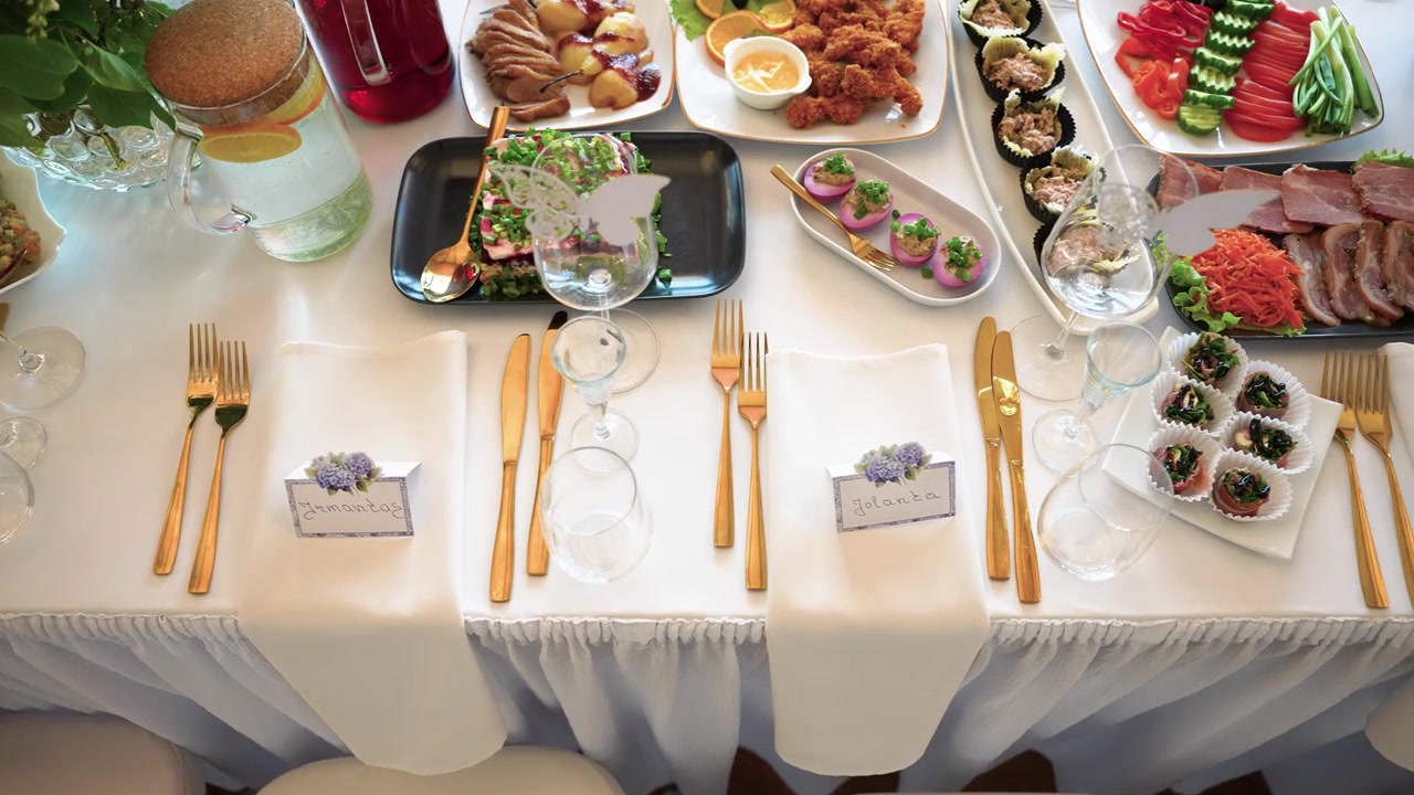 Overhead view of a beautifully set table with gourmet appetizers, gold cutlery, white linens, and place cards, bathed in soft natural lighting. Perfect for upscale celebration footage.