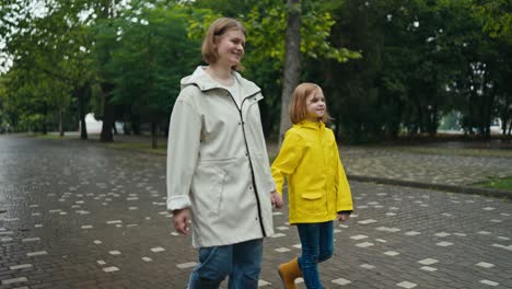 a happy blonde woman in a white jacket walks hand in hand with her teenage daughter in a yellow jacket walking in the park after the rain