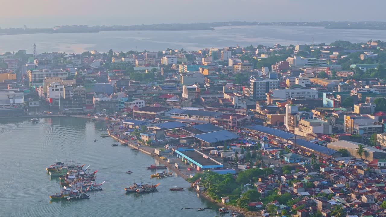 Drone descends over Tacloban City, revealing busy waterfront and downtown buildings at golden hour. Warm light and soft haze frame the scene.