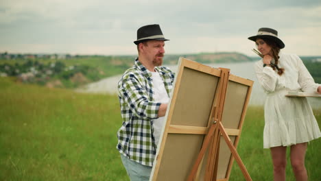 a painter wearing a black hat and plaid shirt is setting up a canvas on an easel in a grassy field. a woman in a white dress stands nearby, smiling as she watches the process