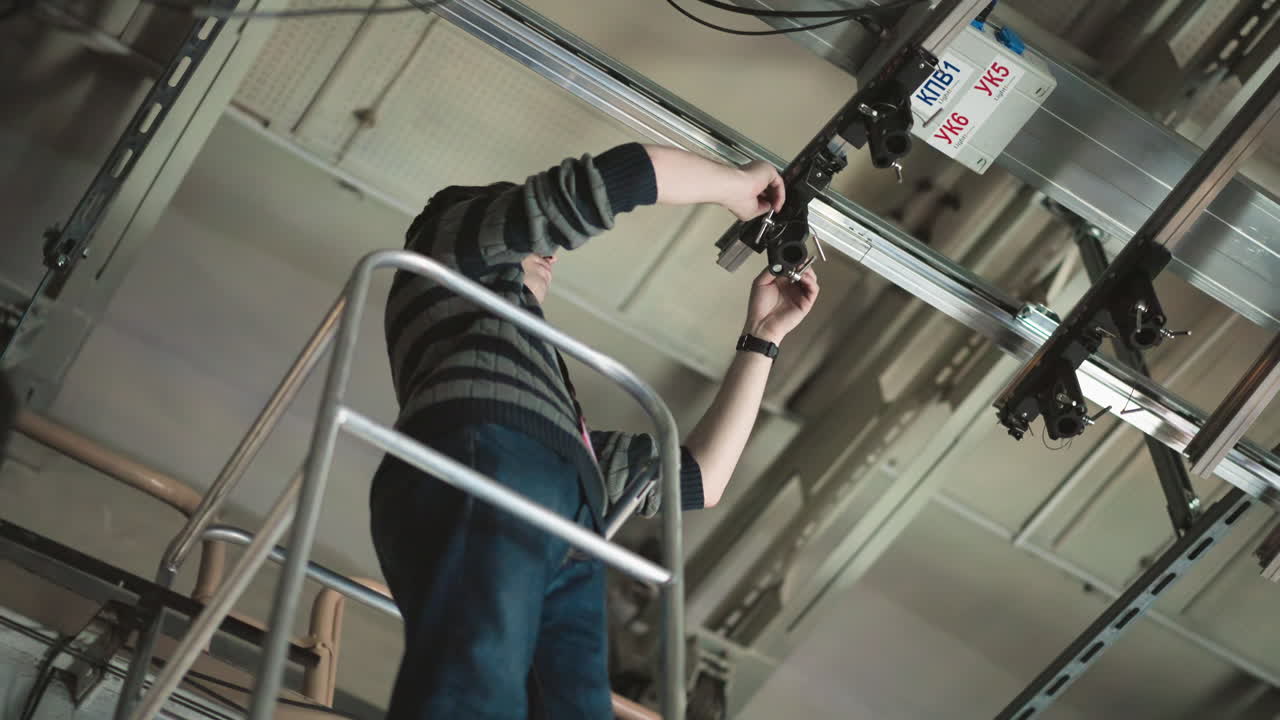 Person working at height, standing on ladder, adjusting camera or lighting equipment mounted on ceiling grid, cables visible, wearing sweater, maintenance or installation task in industrial setting