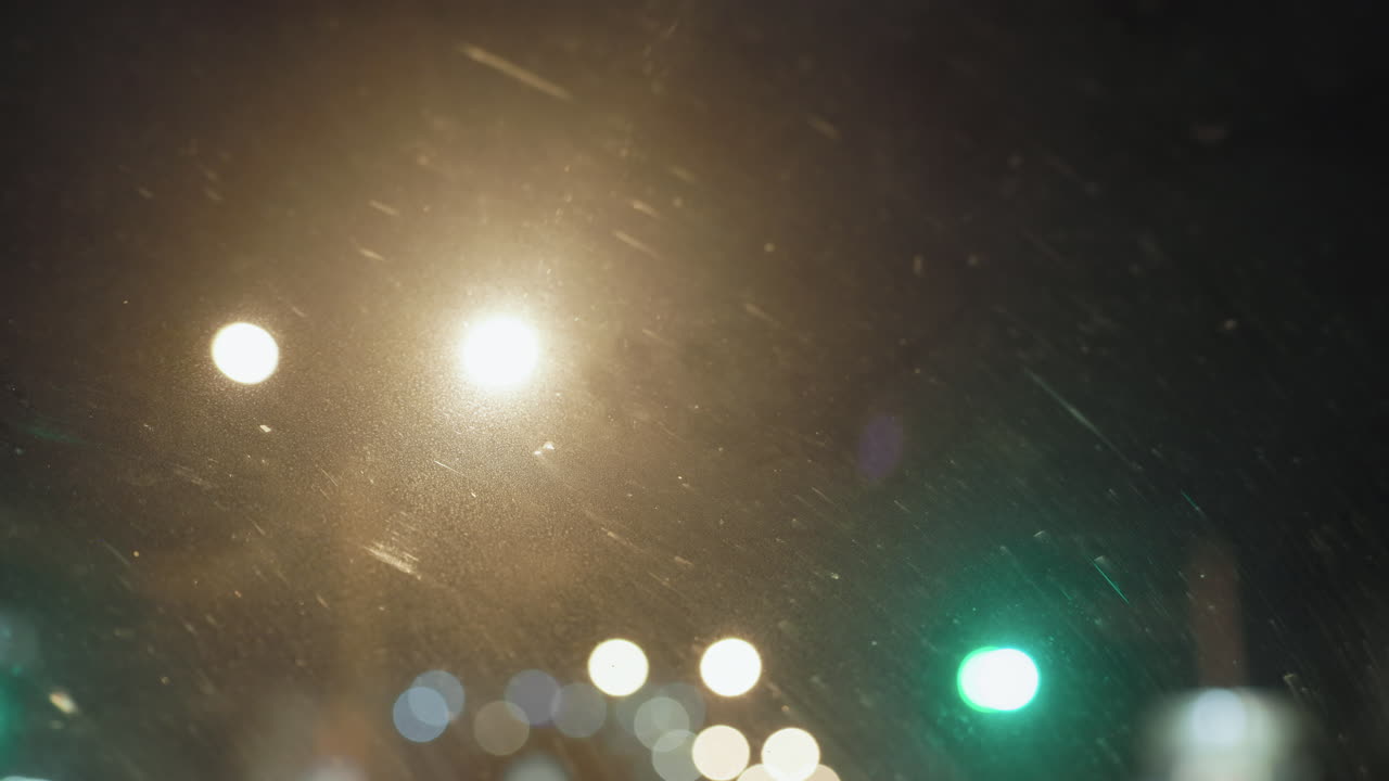 Bokeh light seen through windscreen of moving car at night, bright orbs scattering across dusty glass, traffic signals shining in distance, creating dreamy atmosphere of urban street environment